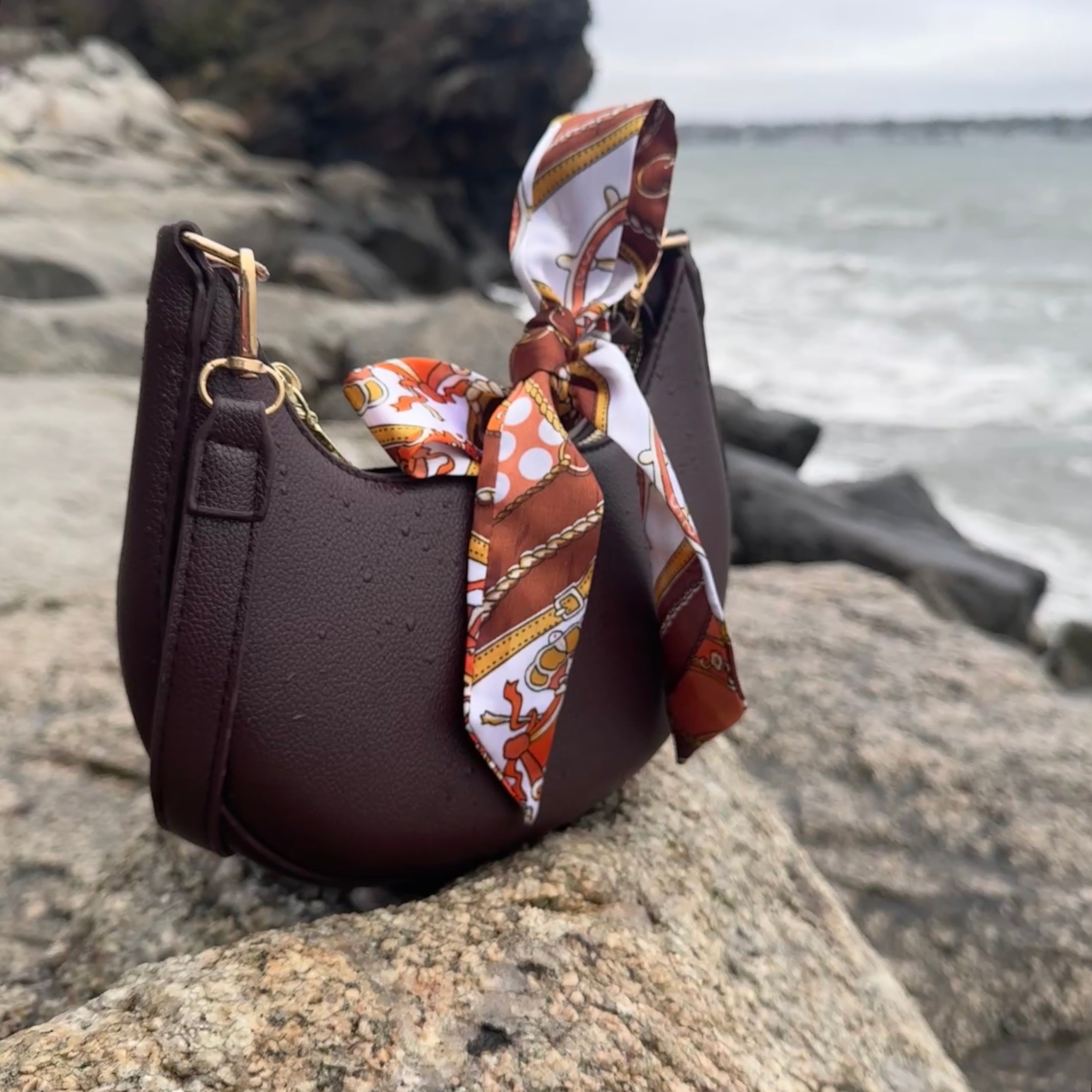 Bordiyari Half Moon Shoulder Bag in brown with ribbon accessory on a rock by the Newport cliffs.